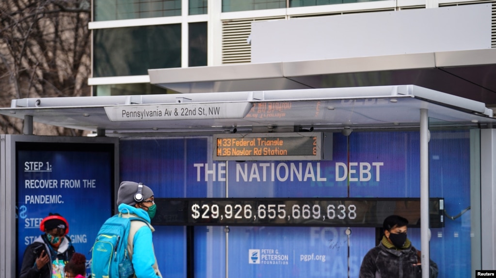 Personas con mascarillas esperan en una parada de autobús, donde se ve un cartel que muestra de la deuda nacional actual en medio de la pandemia de COVID-19 en Washington, el 31 de enero de 2022.