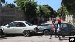 Pejalan kaki berjalan melewati kendaraan yang berfungsi sebagai barikade yang didirikan warga untuk mencegah anggota geng memasuki lingkungan mereka, di pusat kota Port-au-Prince, Haiti, Rabu, 13 November 2024. (Foto: AP)