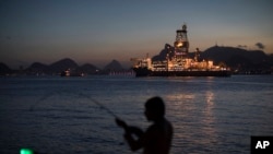 FILE - In this March 28, 2018 photo, a man holds his fishing rod, carrying an illuminated bobber, on the shore of Guanabara Bay where Petrobras operates its ultra-deep drilling vessel Laguna Star NS-44, in Niteroi, Brazil.