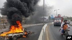 A family on a motorbike rides past a burning vehicle set ablaze by protesters in Karachi , July 14, 2011