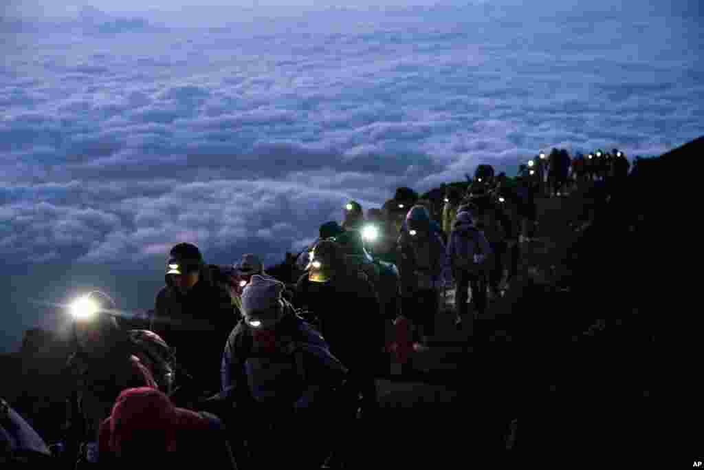 A group of hikers climbs to the top of Mount Fuji, Japan, just before sunrise as clouds hang below the summit.
