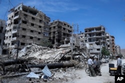 FILE - In this April 16, 2018, file photo, people stand in front of damaged buildings, in the town of Douma, the site of a suspected chemical weapons attack, near Damascus, Syria.