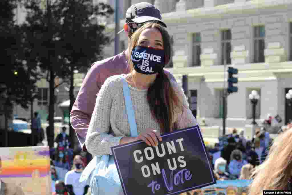 Uma mulher usa uma máscara a dizer "A ciência é real", com um cartaz a dizer "conta conosco para votar". Marcha das Mulheres 17 de outubro 2020