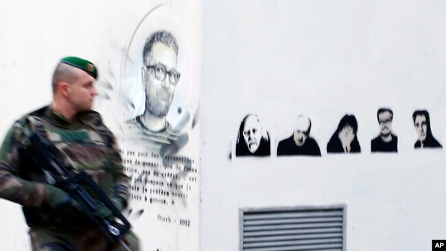 Một binh sĩ Pháp tuần tiễu bên cạnh một bức tranh vẽ chân dung các họa sĩ biếm họa bị giết, Charb (trái), Honore, Wolinski, Cabu, Charb và Tignous bên ngoài tòa soạn cũ của Charlie Hebdo, 1 năm sau cuộc tấn công ở Paris, ảnh chụp ngày 7/1/2016. (AP Photo/Francois Mori)