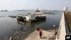 Orang-orang berjalan di dekat tanggul laut raksasa yang digunakan sebagai pembatas untuk mencegah air laut mengalir ke daratan dan menyebabkan banjir di Jakarta, 27 Juli 2019. (Achmad Ibrahim/AP)