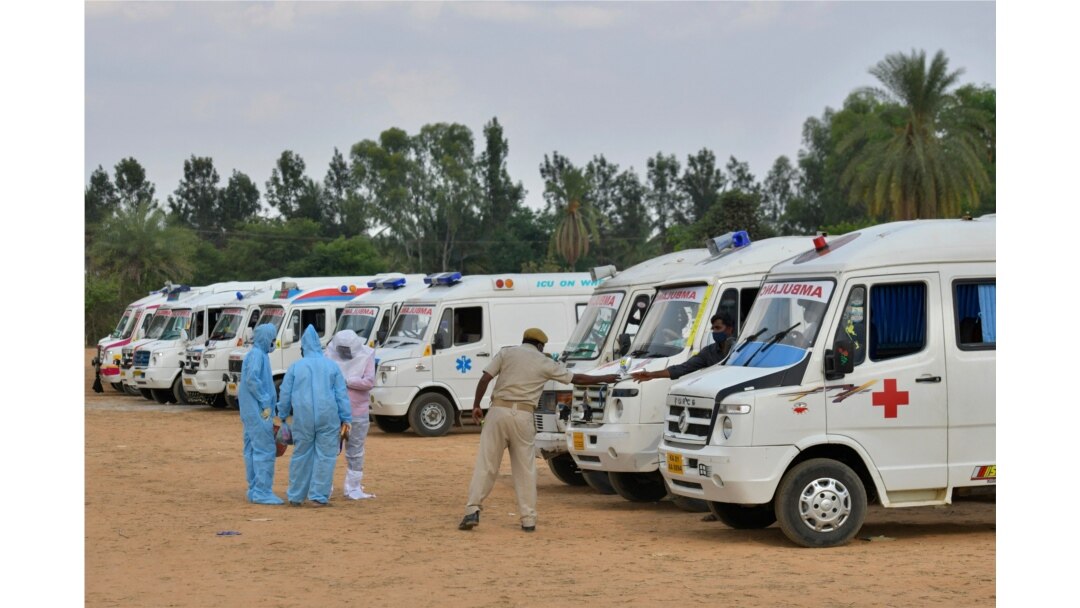 FILE - Family members wearing protective gear stand next to ambulances carrying bodies of COVID victims, at an open air cremation site set up on the outskirts of Bangalore, India, May 8, 2021.