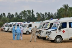 FILE - Family members wearing protective gear stand next to ambulances carrying bodies of COVID victims, at an open air cremation site set up on the outskirts of Bangalore, India, May 8, 2021.