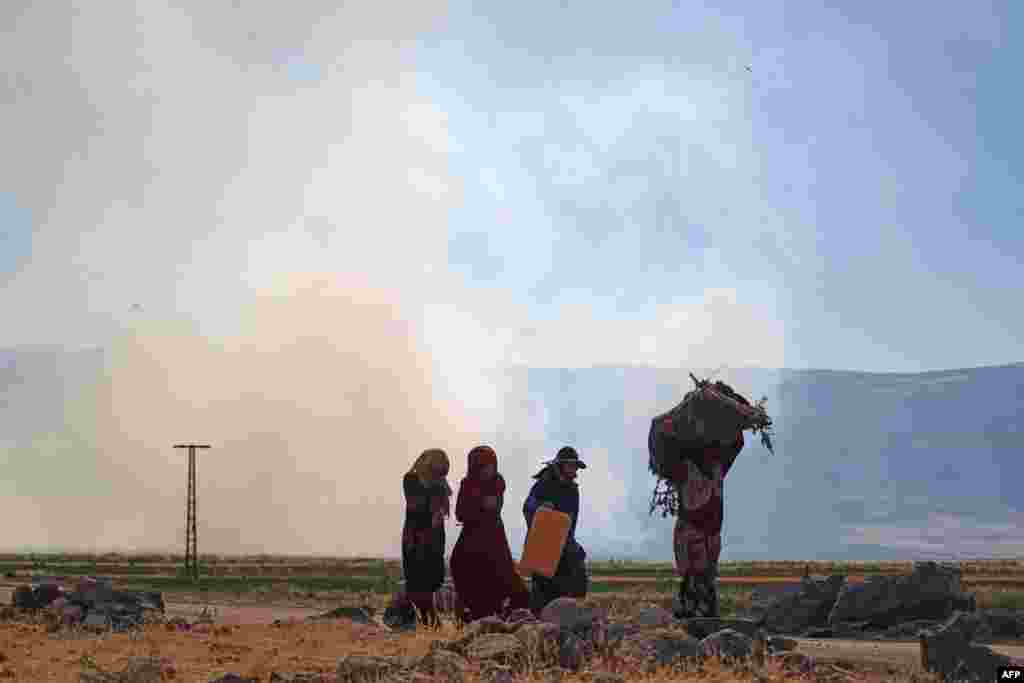 Syrian women walk by as smoke billows in the area of the Syrian village of az-Ziyarah, in the agricultural al-Ghab plain of the Hama province, June 6, 2021, following reported pro-government bombardment.