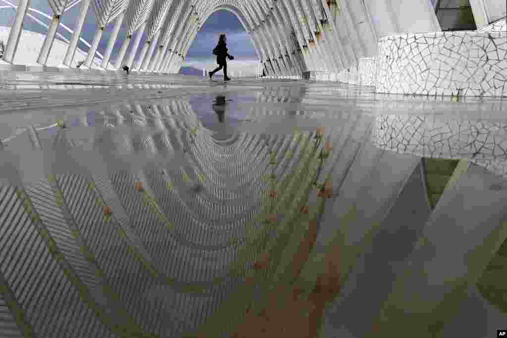 A woman&#39;s image is seen on rain water as she walks inside the modern Agora walkway at the main Olympic complex in Athens, Greece.