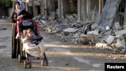FILE - A woman, with her child in a stroller, stands along a damaged street in the besieged area of Homs, Syria.