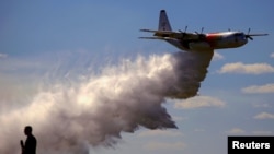 Seorang reporter televisi berdiri di depan Tangki Besar Udara C-130 Hercules yang dikenal dengan nama Thor saat pesawat tersebut menebarkan air 15.000 liter dalam demo oleh Pemadam Kebakaran Desa. 