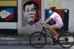 FILE - A woman wearing a protective mask rides her bicycle past an image of Philippine President Rodrigo Duterte in Manila, Philippines, March 20, 2020.