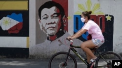 FILE - A woman wearing a protective mask rides her bicycle past an image of Philippine President Rodrigo Duterte in Manila, Philippines, on March 20, 2020. 