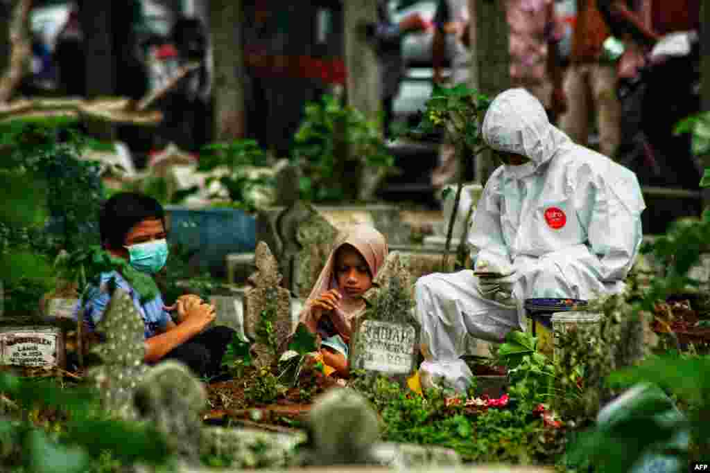 A family member in personal protective equipment sits with two children by the grave of a COVID-19 victim who was just buried, as family members defying health protocols gather nearby at a cemetery in Lhokseumawe, Aceh.