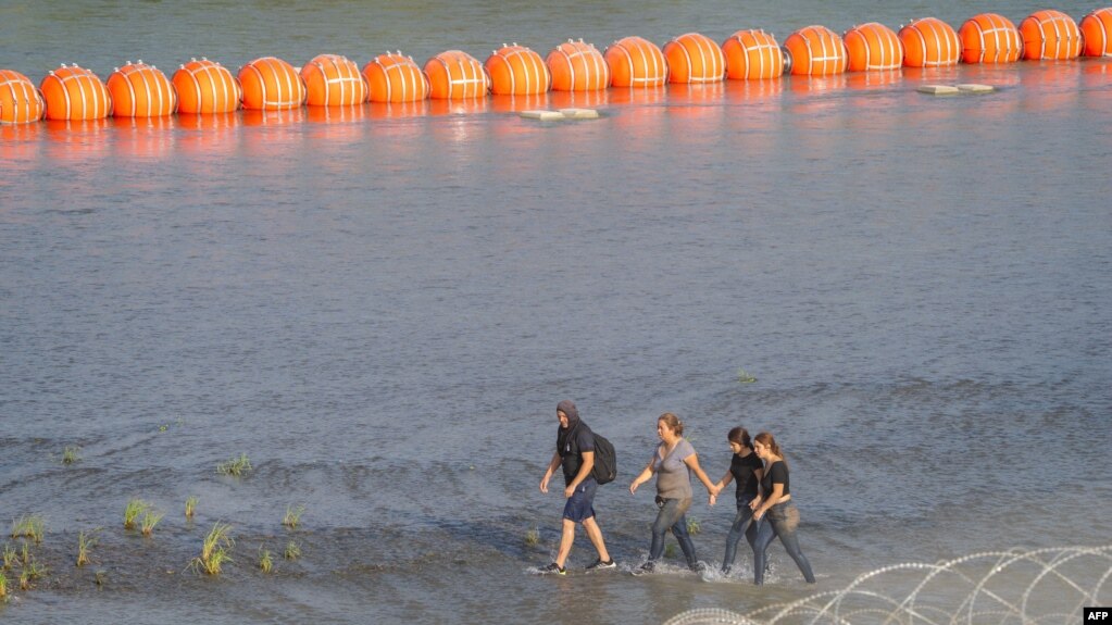 资料照片：移民们在德克萨斯州边境格兰德河边沿着一串橙色浮标行走。（2023年7月17日）(photo:VOA)