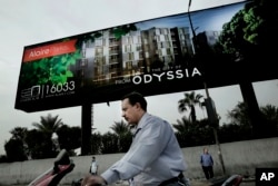 In this Oct. 23, 2018 photo, a man rides his motorbike as others wait for a bus under a billboard promoting a new residential housing compound, in Cairo, Egypt. (AP Photo/Nariman El-Mofty)