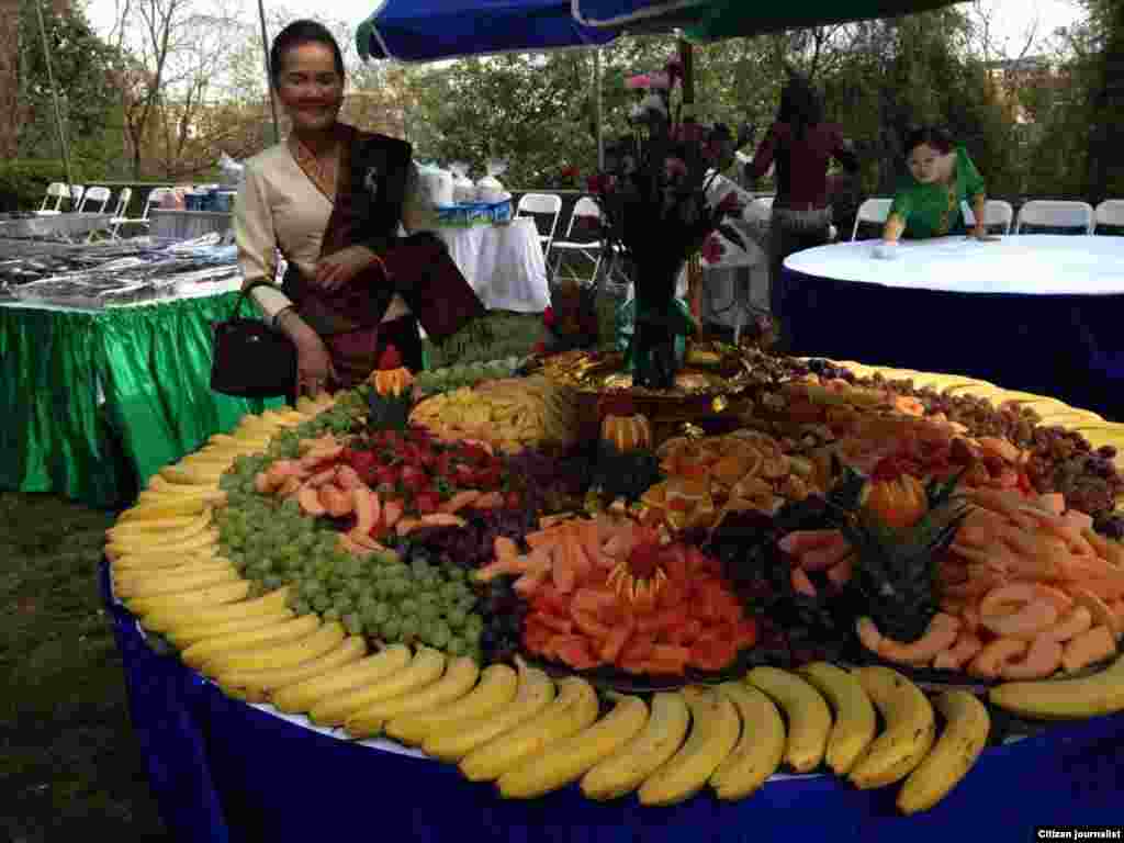 ທ່ານນາງ ສົມດີ ສຸຂະທິວົງ ພັນລະຍາທ່ານທູດແສງ ຢືນຢູ່ຂ້າງໂຕະຜົນລະໄມ້.