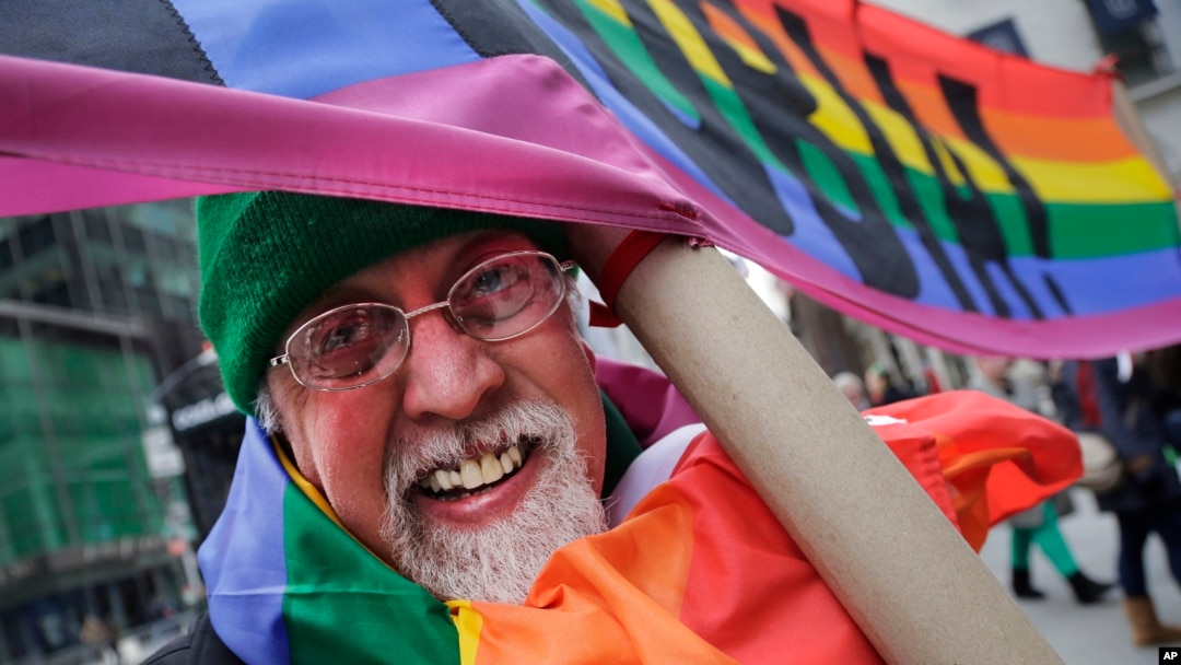 FILE - Artist Gilbert Baker, designer of the Rainbow Flag, is draped with the flag while holding a banner that reads "Boycott Homophobia" before the start of the St. Patrick's Day parade in New York City, March 17, 2014.