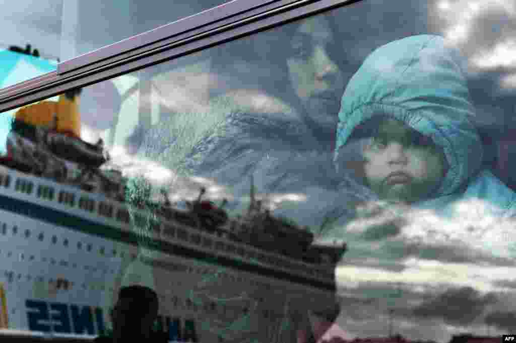 A woman and a child peer from a bus as migrants and refugees disembark from a government chartered ferry, seen in reflection, in the port of Piraeus in Athens, Greece.