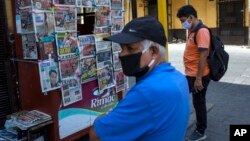 Hombres, con máscaras faciales protectoras, se paran frente a un puesto de periódicos en el centro de Lima, Perú, el sábado 28 de marzo de 2020, durante la segunda semana de un estado de emergencia decretado por el gobierno.