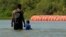 Migrantes caminan junto a grandes boyas que se utilizan como barrera fronteriza flotante en el río Bravo, el 1 de agosto de 2023, en Eagle Pass, Texas.