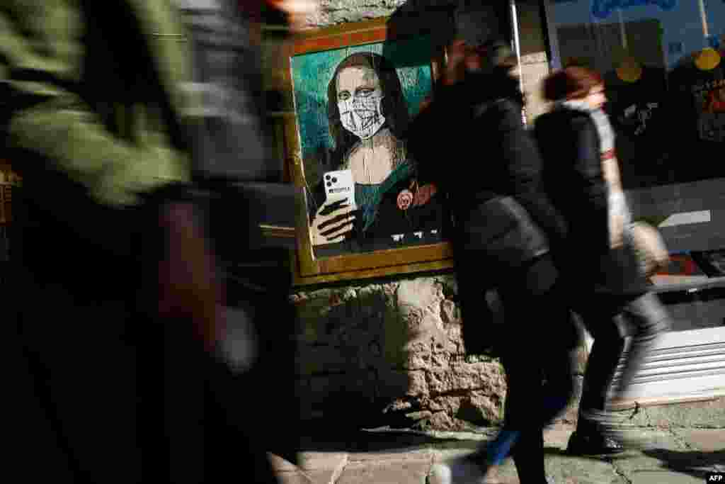 People walk by a poster by Italian urban artist Salvatore Benintende, aka &quot;TVBOY,&quot; depicting Leonardo da Vinci&#39;s Mona Lisa wearing a protective face mask and holding a mobile phone reading &quot;Mobile World Virus&quot; along a street in Barcelona, Spain.