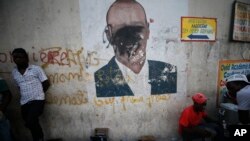 A man shines a client's shoes as another waits for business, in front of a wall painted with an image of President Jovenel Moise, his face obscured by black spray paint, in the Delmas neighborhood of Port-au-Prince, Haiti, Oct. 8, 2019. 