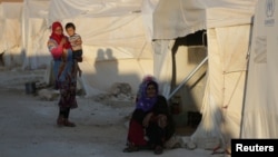 FILE - An internally displaced woman sits outside a tent in Idlib province, Syria, July 30, 2018, as another woman with child looks on. 