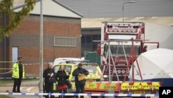 People walk past a police officer at the scene after a truck, seen in rear, was found to contain a large number of dead bodies, in Thurrock, South England, early Wednesday Oct. 23, 2019. Police in southeastern England said that 39 people were found dead W