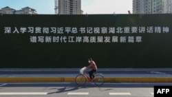 一名戴着口罩的妇女从武汉街头一幅宣传习近平湖北讲话的大标语前骑车而过。（2020年3月10日）