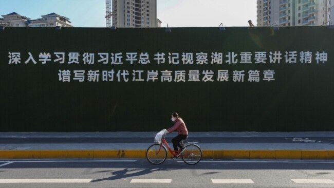 一名戴着口罩的妇女从武汉街头一幅宣传习近平湖北讲话的大标语前骑车而过。（2020年3月18日）
