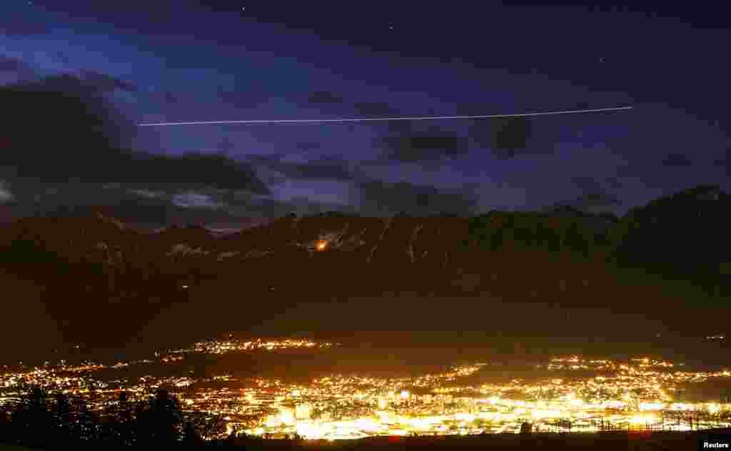 An airplane leaves a light trail as it flies over the snow covered Nordkette mountains behind the city of Innsbruck, Austria.