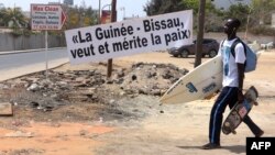 FILE - A young man carrying a skateboard and a surfboard walks past a banner reading "Guinea-Bissau wants and deserves peace," during an Economic Community of West African States (ECOWAS) summit in Dakar, Senegal, June 4, 2016.
