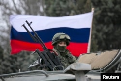 A pro-Russian man (not seen) holds a Russian flag behind an armed servicemen on top of a Russian army vehicle outside a Ukrainian border guard post in the Crimean town of Balaclava, March 1, 2014.
