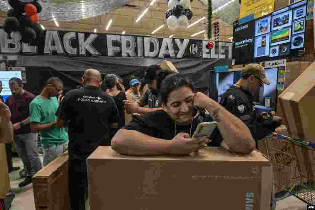 A pistillate   watches her telephone  portion    shoppers bargain  TV sets astatine  a supermarket during a Black Friday merchantability  successful  Sao Paulo connected  Nov. 28, 2024. 