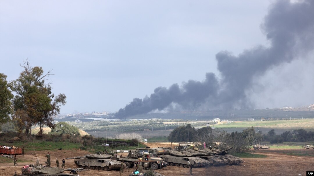 Israeli soldiers stand next to tanks at a position in Israel near the border with the Gaza Strip as smoke billows over the Gaza Strip after Israeli bombardment on January 14, 2024, as the war between Israel and militant group Hamas entered its 100th day.