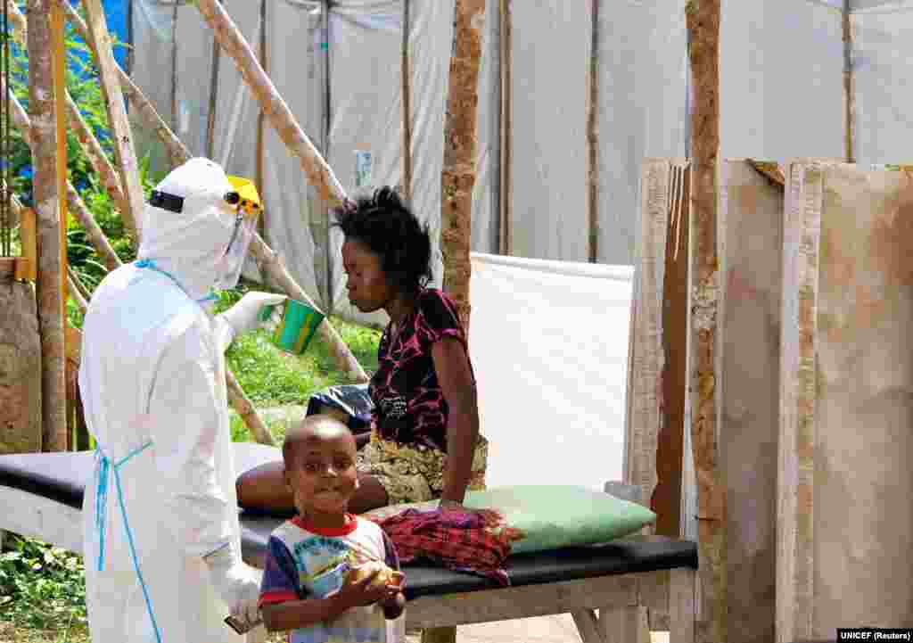 Um técnico de saúde, vestido com equipamento de protecção, oferece água a uma mulher com Ébola num centro de tratamento para pessoas infectadas, enquanto um menino brinca por perto. Hospital Estatal de Kenema, Serra Leoa.