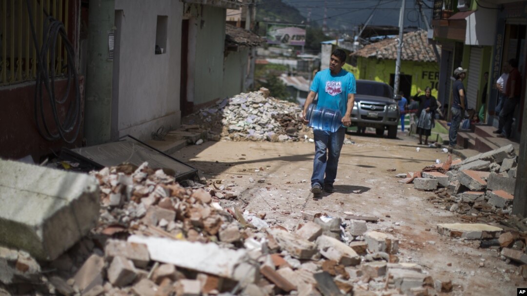 Seorang warga membawa satu galon air melewati jalan yang dipenuhi puing-puing bangunan yang roboh akibat gempa di San Pedro, Guatemala (7/7). (AP/Luis Soto)