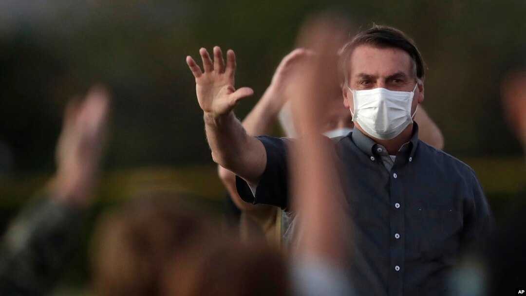 Presiden Brazil Jair Bolsonaro yang terinfeksi Covid-19, mengenakan masker saat melambaikan tangannya kepada para suporternya yang menghadiri upacara penurunan bendera di luar kediaman resminya Istana Alvorada, di Brasilia, Brasil, Senin, 20 Juli 2020. (Foto AP / Eraldo) 