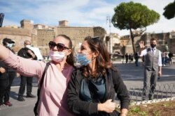 Two women wear a face mask to prevent the spread of COVID-19 as they take a selfie with actor Tom Cruise in the background during a break from shooting Mission Impossible 7, along Rome's Fori Imperiali avenue, Oct. 13, 2020.