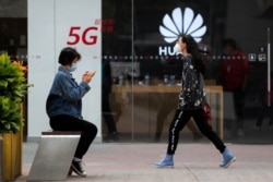 A woman wearing a face mask to help curb the spread of the coronavirus browses her smartphone as a masked woman walks by the Huawei retail shop promoting it 5G network in Beijing Oct. 11, 2020.