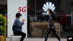 A woman wearing a face mask to help curb the spread of the coronavirus browses her smartphone as a masked woman walks by the Huawei retail shop promoting it 5G network in Beijing Oct. 11, 2020.
