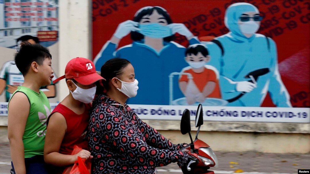 Người dân lái xe ngang qua một biểu ngữ cổ động phòng chống virus corona ở Hà Nội hôm 31/7/2020. Chính phủ Việt Nam đang đàm phán mua vaccine chống COVID-19 từ Mỹ và 3 nước khác trong khi tiếp tục thủ nghiệm lâm sàng để sản xuất vaccine nội địa.