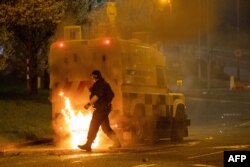 FILE - A police officer walks behind a police vehicle with flames leaping up after violence broke out in Newtownabbey, north of Belfast, in Northern Ireland, April 3, 2021.