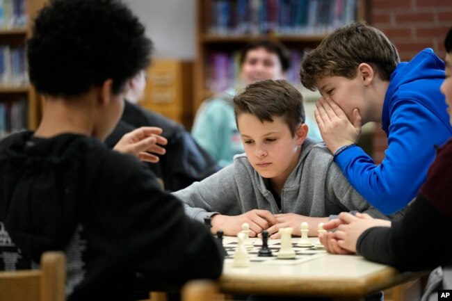 Owen Isenhour whispers advice to Eli Marquis during a Reeds Brook Middle School after-school chess practice, Tuesday, April 25, 2023, in Hampden, Maine. Part-time chess coach and full-time custodian David Bishop led his elementary and middle school teams to state championship titles this year. (AP Photo/Robert F. Bukaty)