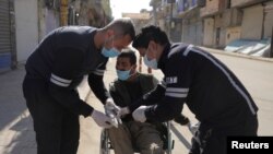 Des agents de santé aident un homme en fauteuil roulant à enfiler ses gants pour empêcher la propagation du coronavirus à Qamishli, Syrie, le 23 mars 2020.
