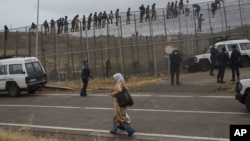 FILE - A woman walks past Sub-Saharan migrants sitting on top of a metallic fence that divides Morocco and the Spanish enclave of Melilla, May 17, 2014. 