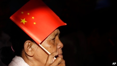 In this photo taken on July 2, 2018, a woman uses a Chinese national flag to protect herself from the sun as she listens to PM Hun Sen who delivering his speech during an inauguration ceremony of a sky bridge in Phnom Penh, Cambodia.