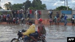 A group of people walk in a partially flooded street by the rains, while another persons try to walk over the water, holding a grating, in the Haitian capital Port-au-Prince, on May 2, 2017.
