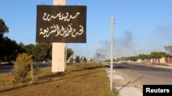 A sign which reads in Arabic, "The city of Sirte, under the shadow of Sharia" is seen as smoke rises in the background while forces aligned with Libya's new unity government advance on the eastern and southern outskirts of the Islamic State stronghold of